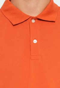 Oranssi polo-paita, joka on valmistettu teksturoidusta kankaasta. Paitassa on kaulus ja kolme valkoista nappia kaula-aukossa. Yksinkertainen, yksivärinen muotoilu, ilman kuvioita.
