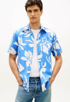 Jeune homme portant une chemise à manches courtes bleue et blanche à motifs floraux sur un t-shirt blanc avec un jean bleu, debout les mains dans les poches.