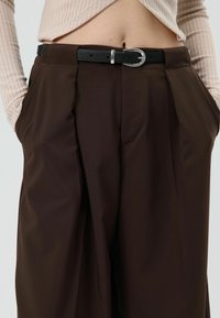 Bruine geplooide broek met een hoge taille, zijzakken en een zwarte leren riem met een zilveren gesp. Getextureerde stof met een gladde afwerking.