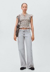 Top floreale corto con maniche a volant, abbinato a jeans larghi grigi chiari e scarpe slip-on nere. Il modello tiene in mano una borsa grigia.