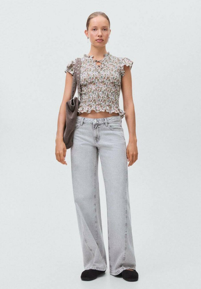Top floreale corto con maniche a volant, abbinato a jeans larghi grigi chiari e scarpe slip-on nere. Il modello tiene in mano una borsa grigia.