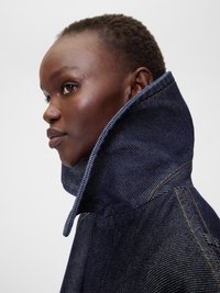 Denim jaka ar augstu apkakli, tumši zila krāsa, redzamā svītru tekstūra un kontrastējošas dzeltenas šuvju detaļas.