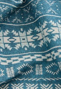 Tyrkisblåt stof med indviklede hvide geometriske mønstre, herunder trekanter, linjer og cirkler, med en blød tekstur og glat udseende.