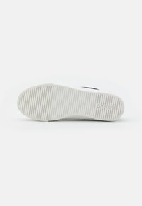Suela de goma blanca de una zapatilla, con ranuras horizontales para tracción y un logotipo grabado cerca del talón. Textura de superficie lisa.