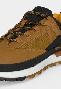 Chaussure de randonnée marron et noire avec des lacets jaunes et noirs, tige en tissu texturé, semelle intermédiaire blanche épaisse et semelle extérieure en caoutchouc robuste.