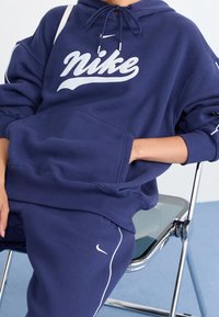 Námořnická mikina s kapucí s bílým logem "Nike", klokáním kapsou a nastavitelnými šňůrkami. Kombinována s odpovídajícími námořnickými tepláky a židlí.