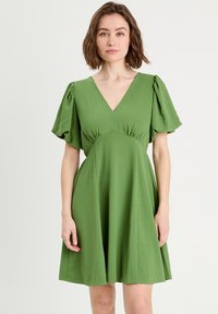 Robe verte en tissu doux, avec un décolleté en v, des manches bouffantes et un corsage ajusté qui s'évasent en jupe.