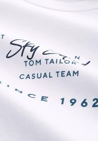 Hvid T-shirt med blå trykt tekst, der læser "Sky Team Tom Tailor Casual Team Siden 1962." Blødt bomuldsstof, casual pasform, glat tekstur.