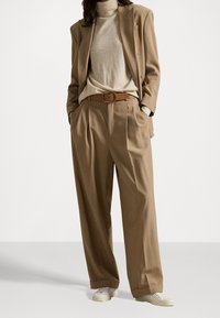 Costume beige avec un blazer ajusté et un pantalon à jambes larges, associé à un col roulé crème et une ceinture marron. Des baskets blanches complètent le look.