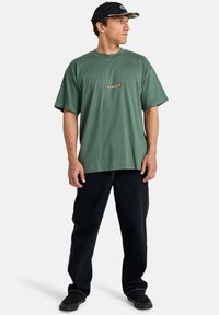 Green cotton t-shirt with a loose fit, featuring a small embroidered logo on the chest. Paired with black pants and a black cap.