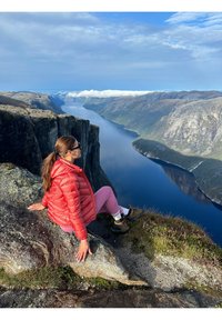 Czerwona kurtka puchowa, różowe legginsy, brązowe buty trekkingowe. Osoba siedzi na skalnej półce z widokiem na rzekę i góry pod niebieskim niebem.