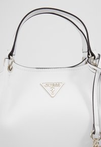 Sac à main blanc avec des accents noirs, fabriqué en matériau lisse. Il présente un logo triangulaire en argent et des poignées doubles solides pour le transport.