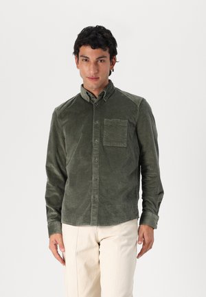Olive green corduroy shirt with a button-down collar, single chest pocket, and vertical ribbed texture. Worn over cream-colored trousers.