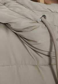 Gepolsterte beige Jacke mit hohem Kragen, verstellbarem Kordelzug mit Metallösen und weicher, glatter Textur.