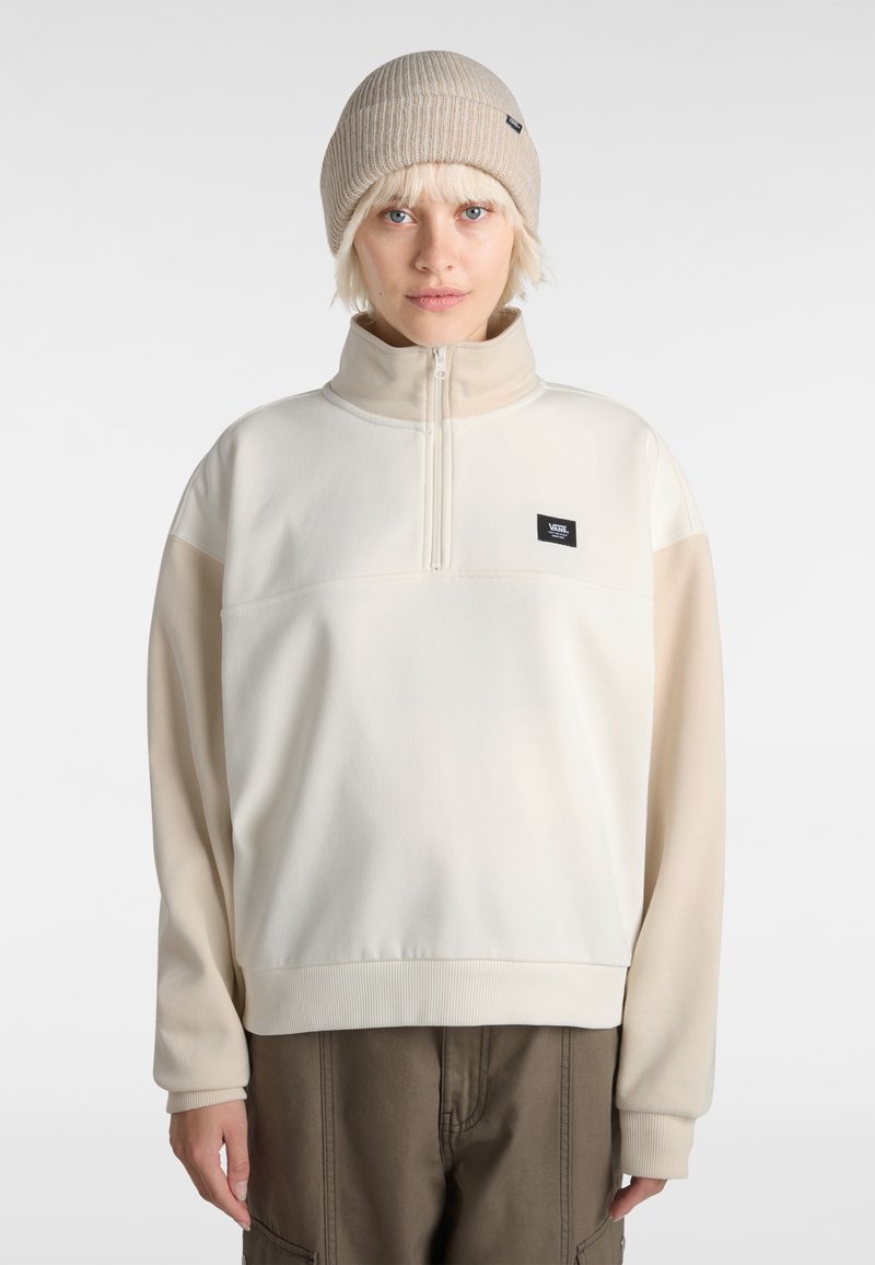 Pull avec col zippé à mi-hauteur en polaire crème clair, manches longues et texture douce ; présente un petit patch logo noir sur la poitrine.