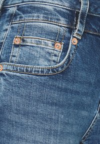 Calças de ganga azul com tecido texturizado, apresentando costuras amarelas contrastantes, rebites de cor cobre e um design clássico de cinco bolsos.