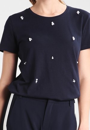 Granatowa bluzka z krótkim rękawem z małymi białymi gronami perełek rozmieszczonymi z przodu, noszona z granatowymi spodniami z białymi paskami z boku.