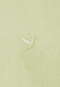 Tejido verde claro con un diseño bordado de un colibrí en un tono ligeramente más oscuro. La textura es suave y tejida.