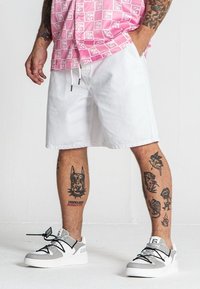 Pantaloni corti bianchi con vita elasticizzata, abbinati a una camicia rosa a maniche corte con fantasia. Sneakers grigie e bianche con dettagli neri. Gambe tatuate visibili.