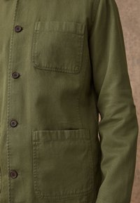 Veste vert olive avec boutons marron, une poche poitrine à gauche et une plus grande poche rectangulaire en dessous sur le côté droit.