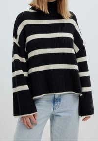 Pull rayé noir et crème avec une texture côtelée, un col montant et des manches larges, associé à un jean bleu clair.