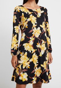 Vestido negro con patrón floral amarillo, mangas largas con puños elásticos y cuello redondo. Confeccionado en una tela suave y lisa.