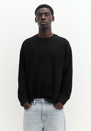 Un homme porte une chemise noire à manches longues et un jean bleu clair. Il se tient les mains dans les poches devant un fond blanc uni.