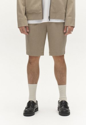 Uomo che indossa pantaloncini beige, giacca abbinata, calzini bianchi e scarpe stringate in pelle nera, in piedi contro uno sfondo uniforme.