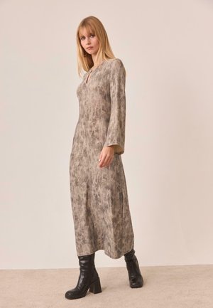 Mujer con un vestido beige de manga larga y largo hasta los tobillos con estampado, y botas negras de tacón, de pie contra un fondo claro y liso.