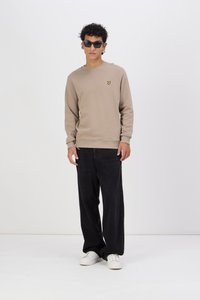 Beige pulóver egy kis sárga logóval a bal mellen, fekete, bő szárú farmernadrággal és fehér sportcipővel párosítva. A modellen fekete napszemüveg van.