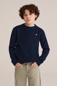 Marinblå stickad tröja med rund halsringning och strukturerat mönster, prydd med en liten vit fågellogo på vänster bröst, bärs över ljusa byxor.