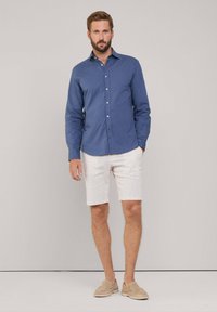 Lichtblauw overhemd met lange mouwen, witte geperforeerde shorts, beige instappers, staande tegen een neutrale achtergrond. Heeft lange mouwen en een kraag.