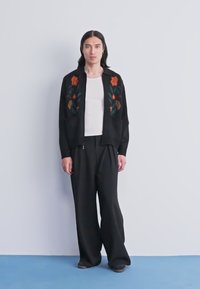 Asmuo su ilgais tamsiais plaukais stovi prieš paprastą sieną, apsirengęs juodu švarku su gėlių raštais, balta palaidine ir plačiomis juodomis kelnėmis.