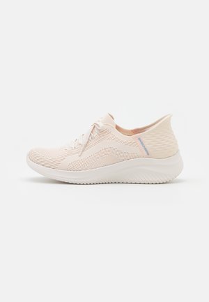 Lyse beige strikkede atletiske sko med snørelukning foran og tekstureret høj hælkant på en hvid sål.