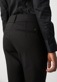 Pantaloni neri su misura con una texture liscia, dotati di una singola tasca posteriore e un dettaglio del logo minimalista. La vita è elegante e piatta.