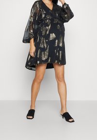 Svart klänning med V-ringning och genomskinliga klockärmar, prydd med metalliska guldfärgade mönster. Bärs med svarta öppna muletofflor.