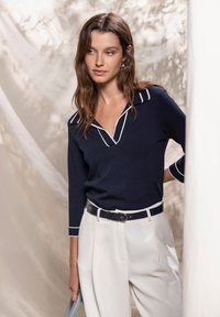 Pull en maille bleu marine avec passepoil blanc, col en V et manches trois-quarts, associé à un pantalon blanc taille haute et une ceinture assortie.