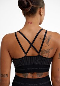 Svart sportbh för aktiviteter med korslagda band, ribbade detaljer och logotyp nederst, bärs av en modell med tatueringar.