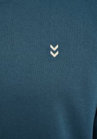 Turkusowy materiał bluzy z gładką fakturą. Charakteryzuje się małym, białym logo w kształcie zygzaka wyszytym po lewej stronie. Prosty, nowoczesny design.