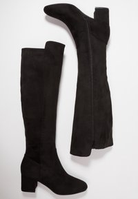 Bottes noires en daim montantes jusqu'au genou avec un talon empilé, dotées d'une fermeture éclair sur le côté et d'une texture lisse, conçues pour une silhouette ajustée.
