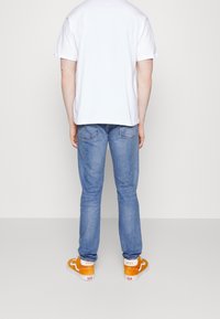 Camiseta blanca, jeans de mezclilla azul clarito y zapatillas Vans naranjas. Diseño visible en la parte posterior con detalles de costura y bolsillos traseros en los jeans.