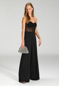 Mustashoudan mustahulben bodice, tapaş ve geniş paça pantolonlar, zımbalı bir çanta ile kombinlenmiş siyah tulum.