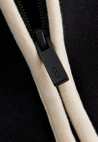 Tirette de fermeture éclair noire avec la lettre « B » attachée à un tissu noir avec un bord en tissu beige de chaque côté de la fermeture éclair.