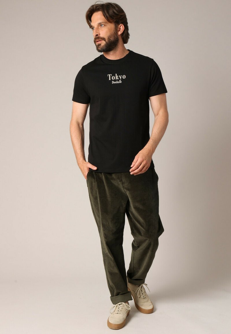 Man met een baard, gekleed in een zwart "Tokyo Deluxe" T-shirt, olijf groene corduroy broek en beige sneakers, staande en kijkend naar de zijkant.