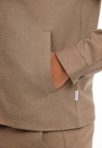 Brun sweatshirt med et struktureret stof. Funktioner inkluderer en forlomma og et mærke på siden, samt en detalje med manchetærmer.