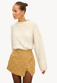 Suéter esponjoso crema con cuello redondo combinado con una falda mostaza con bordado floral blanco y diseño cruzado.