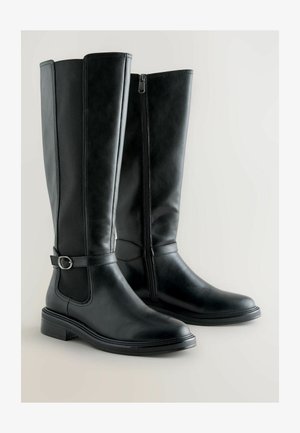 Bottes en cuir noir hauteur genou avec une finition lisse, fermeture éclair sur le côté et une sangle décorative avec une boucle en métal au niveau de la cheville.