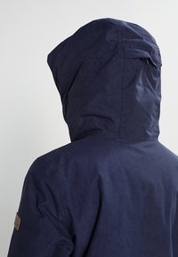Personne portant une veste à capuche bleu foncé vue de dos, montrant le dos et les détails de la capuche sur un fond neutre.
