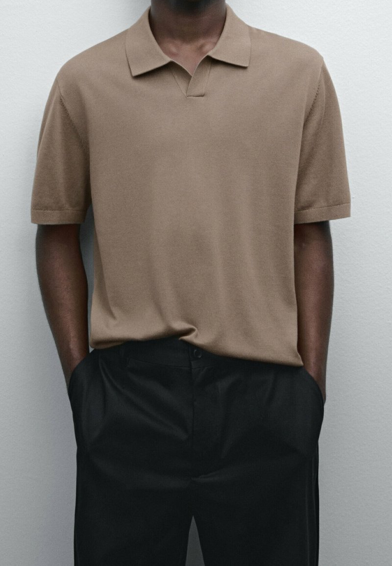 Homme portant un polo à manches courtes beige rentré dans un pantalon noir taille haute, les mains dans les poches, debout contre un mur uni.