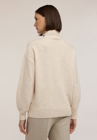 Kobieta z krótkimi brązowymi włosami w beżowym swetrze z golfem i jasnobrązowych spodniach, widoczna z tyłu na jednolitym tle.